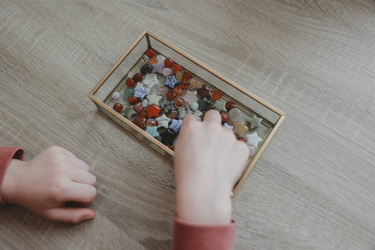 Kinderhanden kiezen een steentje uit een glazen doosje vol gekleurde stenen en sterretjes.